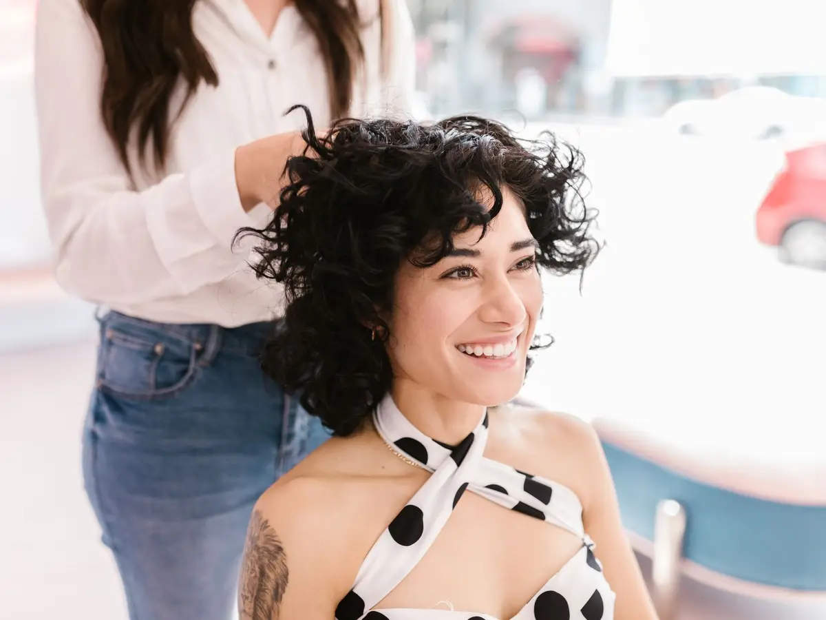 Cliente sorrindo enquanto a cabeleireira finaliza os cachos dela, num salão claro