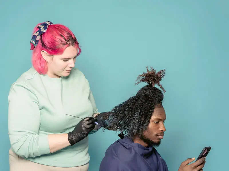 Cliente olhando o celular enquanto a cabeleireira trabalha no cabelo dele